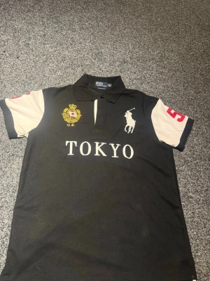Svart Tokyo polo från Polo Ralph Lauren - Cool svart polo från Polo Ralph Lauren med vita ärmar och stora broderade detaljer. Framsidan har TOKYO-tryck, emblem och den klassiska hästloggan. Röd siffra 5 på ärm och rygg. Perfekt för dig som gillar sportig stil.