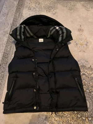 Svart pufferjacka från Burberry - Säljer en svart pufferjacka från Burberry med klassiskt rutigt foder i huvan och dragkedjor på fickorna. Jackan har en stor huva och tydlig Burberry-logga på ärmen. Perfekt för kalla vinterdagar och riktigt snygg streetstil.