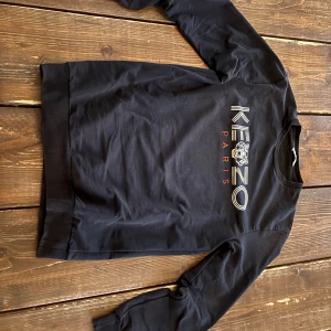 Svart sweatshirt från Kenzo Paris - Säljer en svart sweatshirt från Kenzo Paris med stort logotryck och tigeremblem på bröstet. Tröjan har rund halsringning, långa ärmar och ribbade muddar vid ärmslut och nederkant. Perfekt för dig som gillar streetwear och vill ha något stilrent.