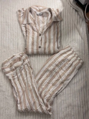 Randig pyjamas  - Säljer en superchill pyjamas i beige och vit med breda ränder. Setet har skjortkrage, knappar framtill och långa ärmar samt byxor med resår i midjan. Tillverkad i ett luftigt linnematerial som känns skönt mot huden. S/M 