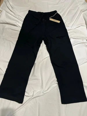 Svarta Essentials mjukisbyxor - Säljer ett par svarta mjukisbyxor från Fear of God Essentials med dragsko i midjan och tryckt logga på benet. Byxorna har raka ben och är gjorda i mjukt bomullsmaterial. Perfekta för en chill och avslappnad stil.