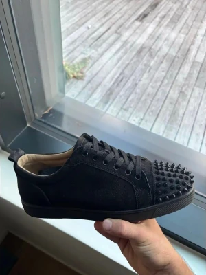  Louboutin svarta size 44 - Säljer ett par svarta sneakers från Christian Louboutin med mattsvarta nitar på tån. Skorna har en låg siluett, snörning och är gjorda i mocka med läderdetaljer. Insidan är beige och sulan är svart. Perfekta för dig som vill sticka ut med en edgy look. Size 44 passar även 43! Använder knappt vill få bort dem asap!