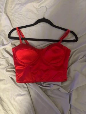 Röd topp - Snygg röd topp i glansigt material med vadderade kupor och justerbara axelband. Toppen har en croppad passform och markerade sömmar framtill för extra stöd och form. Köptes från New Yorker. Säljs då den ej passar mig.