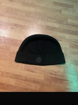 Svart ribbstickad mössa från Moncler - En svart ribbstickad mössa från Moncler med uppvikt kant och Moncler-logotyp framtill. Klassisk och enkel design som passar till många olika stilar. Materialet är mjukt och värmande, perfekt för kalla dagar. För frågar så är det bara och skriva. Mvh✌️