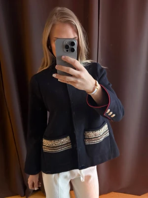 Blå kappa  - Unik marinblå kappa från Maison Cherie med dekorativa broderier och band i guld och silver på fickorna. Kappan har raka ärmar, knappar framtill och snygga detaljer med röda insidor på ärmsluten samt guldknappar. Knappt använd. 