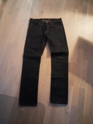 Mörkblå jeans från Dr Denim - Säljer ett par mörkblå jeans från Nudie Jeans med klassisk femficksmodell och kontrastsömmar i orange. Jeansen har rak passform och är tillverkade i kraftig denim. Perfekta för dig som gillar stilrena och tidlösa jeans.