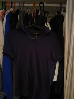 Mörkblå henley från H&M - Säljer en mörkblå henley från H&M i slim fit-modell. Skjortan har korta ärmar, knappar vid halsen och är tillverkad i mjuk bomull. Perfekt för dig som gillar en enkel men stilren look.