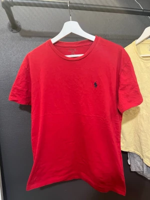 Röd t-shirt från Ralph Lauren - Snygg röd t-shirt från Ralph Lauren med klassisk blå broderad logga på bröstet. T-shirten har rund halsringning och korta ärmar. Tillverkad i mjuk bomull som känns skön mot huden. Perfekt för dig som gillar stilrena och tidlösa plagg.