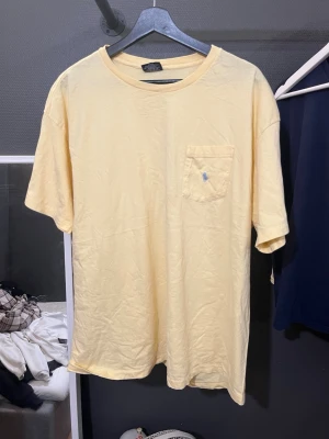 Gul t-shirt från Polo Ralph Lauren - Ljusgul t-shirt från Polo Ralph Lauren med klassisk passform. T-shirten har korta ärmar, rund halsringning och en bröstficka med blå broderad logga. Tillverkad i mjuk bomull som känns skön mot huden.