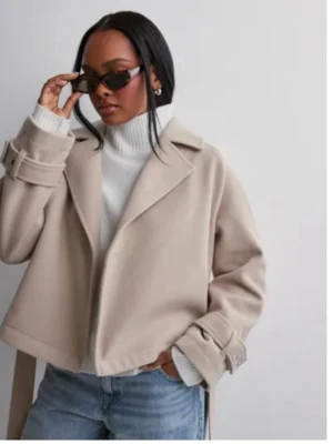 Beige kort kappa med bälte - Snygg beige kappa med breda slag och bälte i midjan. Kappan är kortare i modellen och har dekorativa spännen vid ärmsluten. Perfekt för dig som vill ha en stilren och trendig look. Materialet ser ut att vara mjukt och ull-liknande.