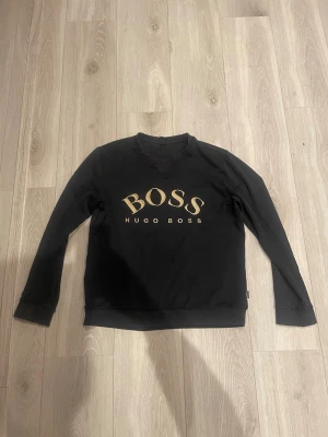 Svart sweatshirt från Hugo Boss - Snygg svart sweatshirt från Hugo Boss med stort guldfärgat BOSS-tryck på bröstet och texten HUGO BOSS under. Tröjan har rund halsringning och långa ärmar. Tillverkad i mjukt material som känns skönt mot huden. Perfekt för en stilren och avslappnad look.