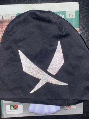 Svart mössa från SOC - Svart mössa med ett stort, stilrent reflextryck framtill. Mössan har en enkel och klassisk form som passar perfekt till kalla dagar. Reflexdetaljen gör att du syns bättre i mörkret. Perfekt accessoar för dig som vill kombinera stil och säkerhet.