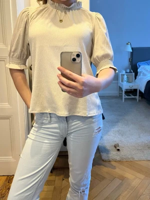 Ljusbeige blus  - Säljer en ljusbeige blus med puffiga trekvartsärmar och rynkad hög hals. Blusen har en lätt glansig yta och avslappnad passform. Perfekt att styla med jeans eller kjol för en trendig look.