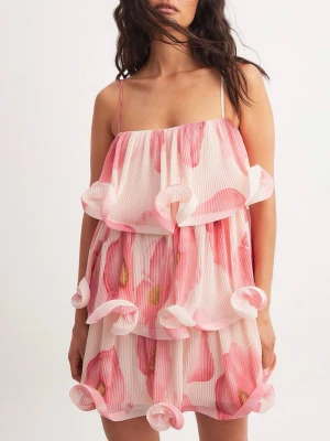 Blommig volangklänning med tunna band - Supersöt kort klänning med volanger och tunna axelband. Klänningen har ett blommigt mönster i rosa och vitt, samt plisserat tyg som ger extra struktur. Perfekt för sommarens alla tillfällen och ger en lekfull vibe.