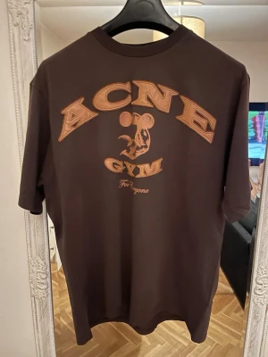 Brun Acne Studios t-shirt med tryck - Cool brun t-shirt från Acne Studios med stort tryck i orange på ryggen där det står 'ACNE GYM' och en illustration av en muskelmus. T-shirten har rund hals och korta ärmar, perfekt för en avslappnad streetstyle-look.