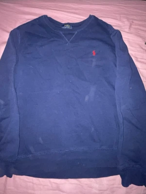 Blå sweatshirt från Polo Ralph Lauren - Säljer en blå sweatshirt från Polo Ralph Lauren med klassisk rund hals och diskret röd logobrodyr på bröstet. Tröjan har långa ärmar och är gjord i mjukt bomullsmaterial. Perfekt för en avslappnad och stilren look.