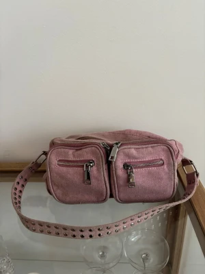 Rosa noella axelväska med nitar  - Säljer en rosa axelväska i canvas med två framfickor och silvriga dragkedjor. Väskan har en bred axelrem dekorerad med nitar fö . Från märket noella