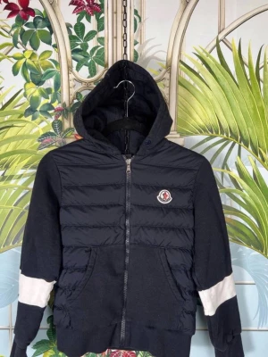Svart Moncler hoodie med dragkedja - Snygg svart hoodie från Moncler med quiltad front och vita detaljer på ärmarna. Dragkedja framtill, två fickor och klassisk Moncler-logga på bröstet. Huva och ribbade muddar ger en sportig och exklusiv look. Perfekt för dig som gillar streetwear och premiumkänsla.