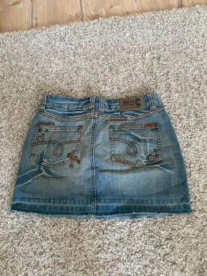 Blå jeanskjol med dekorativa knappt - Säljer en skitsnygg kort jeanskjol med slitna detaljer och dekorativa knappar vid fickorna. Kjolen har klassiska fickor fram och bak, bälteshällor och kontrastsömmar. Finns inte att köpa helt ny och Storleken är 34, pris kan disskuteras💗💗