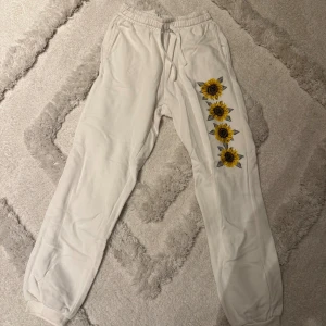 Vita mjukisbyxor med solrosmotiv - Säljer ett par vita mjukisbyxor med resår i midjan och vid bensluten. På ena benet finns fyra stora solrosor tryckta i gult och brunt som ger en unik och färgglad touch. Perfekta för chill dagar hemma eller när du vill ha en skön och avslappnad stil.