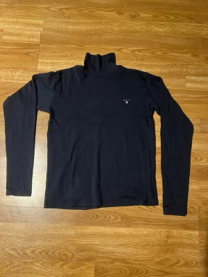 Svart polotröja från Gant - Klassisk svart polotröja från Gant med liten logga på bröstet. Tröjan har lång ärm och är tillverkad i ett mjukt, tunt material som passar perfekt för lager på lager. Enkel och stilren design som funkar till det mesta.