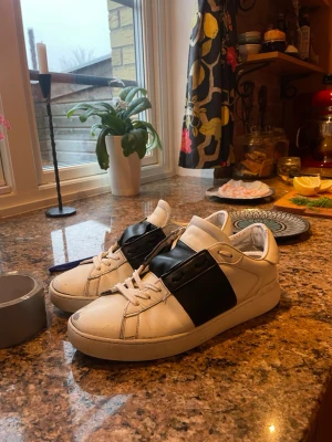Valentino opens  - Säljer ett par vita Valentino sneakers med ett brett svart band över vristen. Skorna har klassisk låg profil, vita snören och detaljer i läder. Perfekta för dig som gillar stilrena men ändå unika sneakers.