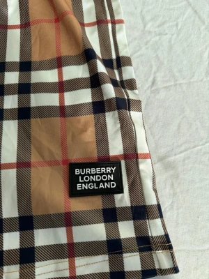Rutig Burberry shorts med snörning - Säljer ett par klassiska Burberry shorts med ikoniskt rutigt mönster i beige, svart, vitt och rött. Shortsen har elastisk midja med svart snörning och en Burberry London England-patch på benet. Perfekta för en trendig och avslappnad stil.