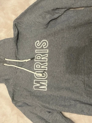 Grå hoodie från Morris - Snygg grå hoodie från Morris med vit broderad logga på bröstet. Hoodien har huva med vita dragsnören och en klassisk känguruficka framtill. Perfekt för en avslappnad och trendig stil.