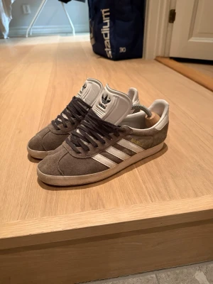 Adidas Gazelle grå sneakers - Snygga Adidas Gazelle sneakers i grå mocka med vita detaljer och klassiska tre ränder på sidorna. Skorna har vit plös med Adidas-logga och guldtext på sidan. Lågt skaft och gummisula ger en clean och tidlös look. Bra skick endast lite smutsiga. Storlek 41-42 