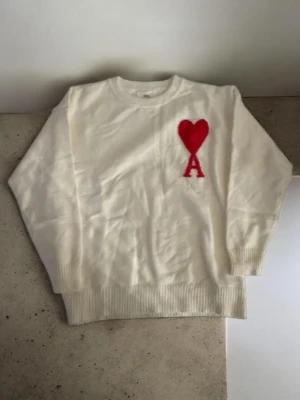 Vit stickad tröja från Ami Paris - Vit stickad tröja från Ami Paris med röd hjärtlogga och bokstaven A på bröstet. Tröjan har rund halsringning, ribbade muddar och är tillverkad i mjukt material. Perfekt för en stilren och trendig look.