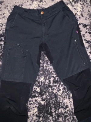 Svarta cargobyxor med dragkedjor - Snygga svarta cargobyxor med flera fickor och dragkedjor, perfekta för en streetwear-look. Byxorna har förstärkta partier vid knäna och en liten brittisk flagga på sidan. Materialet känns slitstarkt och passar till en aktiv vardag. Perfekt för jobb.