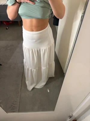 Vit långkjol med volangdetaljer - Säljer en snygg vit långkjol med bred midja och volangdetaljer. Kjolen har en flowy passform och är perfekt för varma dagar. Tillverkad i ett mjukt och lätt material som ger en avslappnad look.