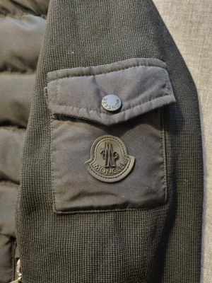 Moncler Cardigan  - Snygg svart dunjacka från Moncler med quiltad front👌, stickade ärmar och ribbade muddar. Jackan har två dragkedjeförsedda fickor fram och en ficka med Moncler-logga på ärmen🔥. Hög krage och stilren design, perfekt för kalla dagar. Skriv för mer info🙂