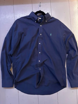 Marinblå skjorta från Ralph Lauren - Klassisk marinblå skjorta från Ralph Lauren med button-down krage och vita knappar. Skjortan har lång ärm och den ikoniska broderade loggan i grönt på bröstet. Tillverkad i mjuk bomull för en bekväm känsla.