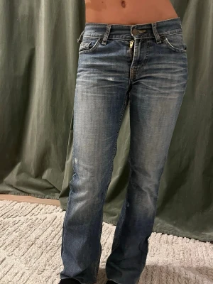 Blå bootcut jeans med slitningar - Snygga blå bootcut jeans med lätt slitna detaljer och klassisk femficksdesign. Jeansen har låg midja, dragkedja och knapp framtill samt markerade sömmar. Perfekta för en avslappnad och trendig look. Dradkedjan är trasig så den går inte att stänga men knappen funkar som vanligt.