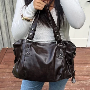 Mörkbrun handväska i pu-läder - Snygg mörkbrun handväska i PU läder med dubbla axelband och silverfärgade detaljer. Väskan har en mjuk, rundad form och dekorativa sömmar framtill. Perfekt storlek för att få plats med allt du behöver och passar till många olika outfits.