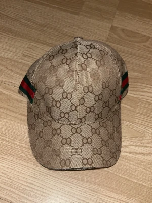 Beige keps från Gucci med GG-mönster - Säljer en beige keps från Gucci med det klassiska GG-monogrammet över hela kepsen. På sidorna finns gröna och röda ränder som ger en ikonisk touch. Justerbar rem baktill med metallspänne. Materialet är canvasliknande och kepsen har böjd skärm. OBS FAKE