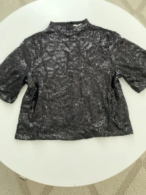 Svart glittrig topp från H&M - ALDRIG använd topp från H&M i storlek XS. Toppen har korta ärmar, hög hals och är täckt av paljetter. Perfekt att ha på fest, firande, nyår, jul eller bara för att se extra fin ut .