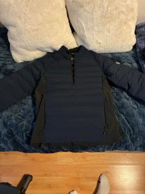 Marinblå vindjacka från Sail Racing - Snygg marinblå vindjacka från Sail Racing med halv dragkedja framtill och två sidofickor med dragkedja. Jackan har en sportig design med vadderade partier och hög krage. Perfekt för blåsiga dagar och en aktiv stil.