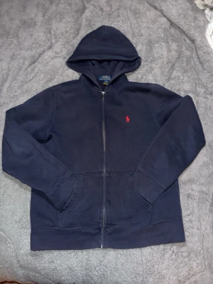 Mörkblå hoodie från Polo Ralph Lauren - Mör sopblå hoodie med dragkedja från Polo Ralph Lauren. Klassisk bro thermad röd logga på bröstet, två fickor framtill och ribbade muddar. Tillverkad i mjuk bomullsblandning och har en stor huva för extra komfort.