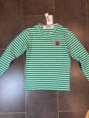 Randig långärmad topp Comme des Garçons - Grön och vit randig långärmad topp från Comme des Garçons PLAY med rund halsringning och det ikoniska röda hjärtat broderat på bröstet. Tillverkad i mjuk bomull som känns skön mot huden. Perfekt för dig som gillar streetstyle och vill sticka ut.