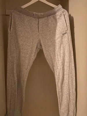 Grå mjukisbyxor från Hugo Boss - Snygga grå mjukisbyxor från Hugo BOSS med vita dragkedjefickor och svart logotyptryck på benet. Byxorna har resår i midjan och mudd vid bensluten. Perfekta för chill dagar och streetwear-stil.