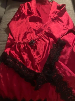 Rött satinnattlinne med spetsdetaljer - Snyggt rött nattlinne i glansigt satinmaterial med svarta spetsdetaljer längs nederkanten och bysten. Har en liten svart rosett framtill och justerbara axelband. Perfekt för en lyxig och bekväm kväll hemma. Med shorts 