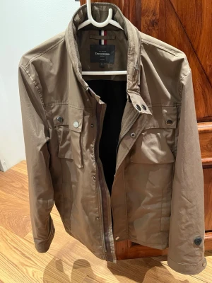 Feild jacket från Dressmann - Snygg brun jacka från Dressmann med ståkrage och flera fickor framtill. Jackan har dragkedja och tryckknappar, samt detaljer vid axlarna. Perfekt för dig som vill ha en stilren och praktisk jacka till vardags.