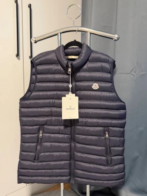  Moncler väst - Snygg mörkblå dunväst från Moncler med horisontella sömmar, hög krage och två dragkedjefickor framtill. Västen har en diskret Moncler-logga på bröstet och stängs med dragkedja. Perfekt för lager-på-lager och stilren look.