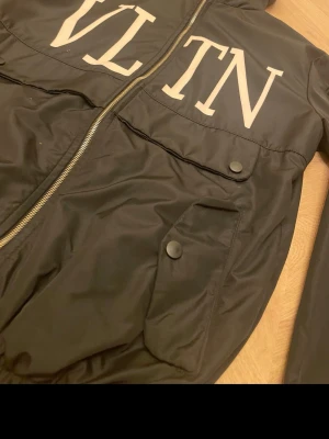 Svart vindjacka från V - Svart vindjacka från V med stor vit V-logga över bröstet. Jackan har huva med dragsko, två stora fickor med lock och tryckknapp samt dragkedja framtill. Materialet är glansigt och lätt, perfekt för blåsiga dagar.