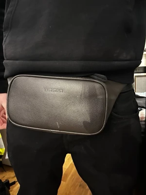 Valentino magväska - Snygg svart magväska från Valentino i slätt konstläder med diskret logga framtill. Väskan har en minimalistisk design och justerbart band som gör den enkel att bära runt midjan eller över axeln. Perfekt för att hålla koll på dina viktigaste prylar.