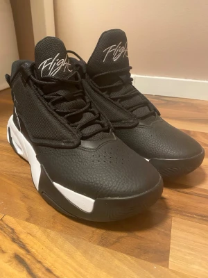 Nike Jordan Max Aura 4 - Säljer ett par svarta Jordan Flight sneakers med högt skaft och vita detaljer på sulan. Skorna har en robust design, snörning framtill och är tillverkade i en kombination av syntetmaterial och textil. Ikonisk Jumpman-logga syns på sulan och Flight-tryck på plösen.