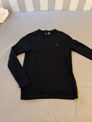 Svart långärmad tröja Polo Ralph Lauren - Klassisk svart långärmad tröja från Polo Ralph Lauren med rund halsringning och den ikoniska röda logotypen broderad på bröstet. Tillverkad i mjuk bomull och har ribbade muddar vid ärmslut och hals. Perfekt för en stilren och enkel look.