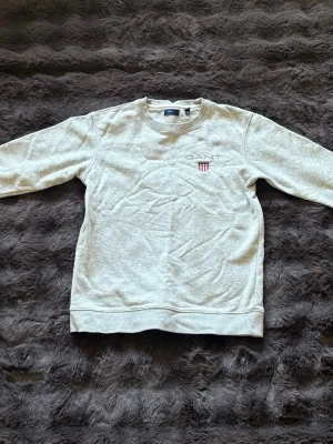Ljusgrå sweatshirt från GANT - Säljer en ljusgrå sweatshirt från GANT med rund halsringning och broderad logga samt sköld på bröstet. Tröjan har långa ärmar och ribbade muddar vid ärmslut och nederkant. Perfekt för en avslappnad och stilren look.